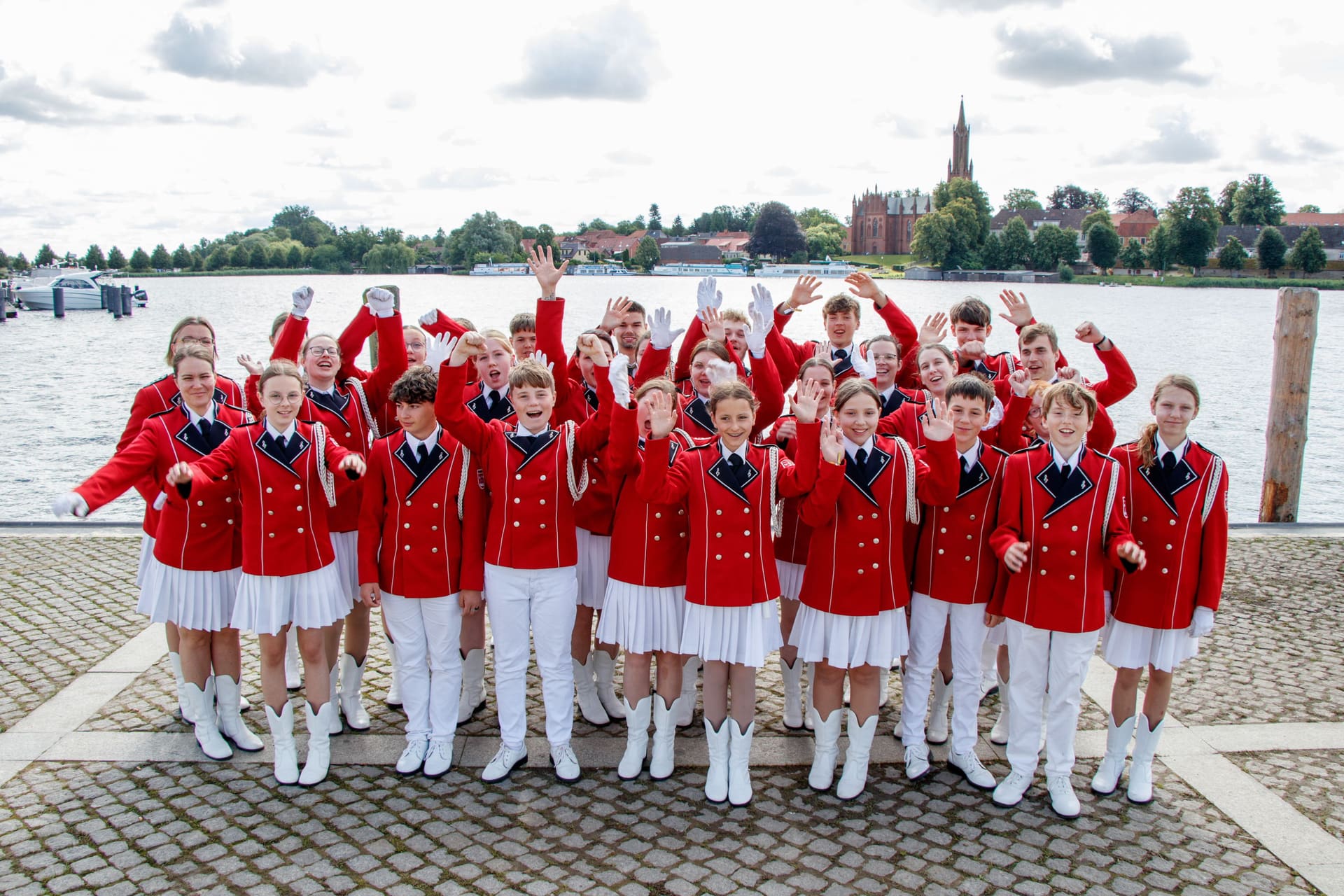 Die Band in roten Jacken und weißen Hosen am Seeufer, alle winken in die Kamera